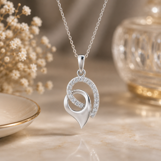 A silver pendant diamond necklace with interlocking curved heart and crescent motifs, one adorned with small clear diamond , displayed on a warm beige surface with soft decorative props in the background.