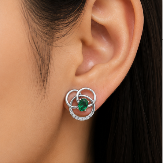 A close-up of a person's ear wearing a silver stud diamond earring with interlocking circular rings, a small oval green gemstone, and a curved row of clear diamond.