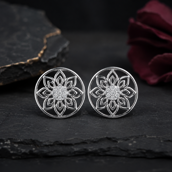 A pair of silver-toned circular stud diamond earrings with an intricate floral mandala filigree design and a sparkling diamond center, displayed on a dark slate surface with a blurred dark rose in the background.