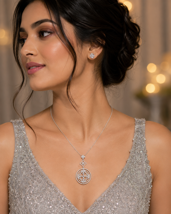 A woman wearing a silver-toned pendant necklace and matching stud earring, with her hair in an updo and a sparkling silver sequined dress against a softly blurred warm background.