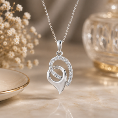 A silver pendant diamond necklace with interlocking curved heart and crescent motifs, one adorned with small clear diamond , displayed on a warm beige surface with soft decorative props in the background.