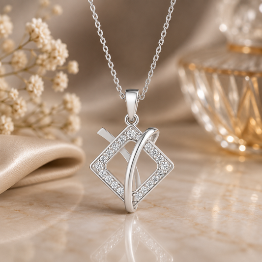 A silver pendant necklace with a diamond-shaped frame set with small clear diamonds and an intertwined curved bar, displayed on a beige marble surface with soft cream flowers and a glass bottle in the background.