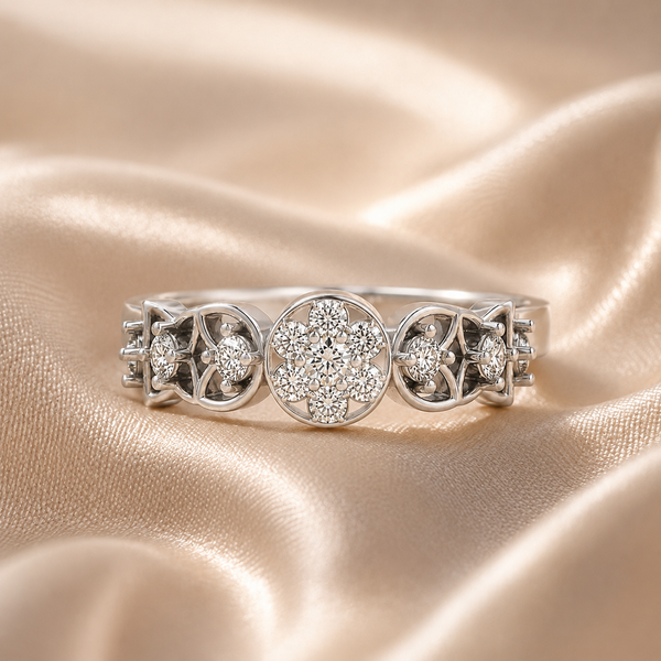 A silver three-stone ring featuring a central round cluster of clear stones flanked by two smaller matching clusters, displayed on a draped champagne satin fabric.