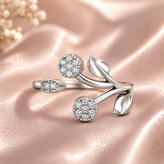 A silver-toned open ring designed like stylized leaves and buds, set with sparkling clear pavé gemstones, displayed on a draped pink satin fabric background.