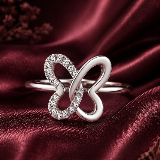 A silver ring featuring a double infinity symbol design, with one loop paved with small clear stones and the other loop polished silver, resting on a draped dark red satin fabric.