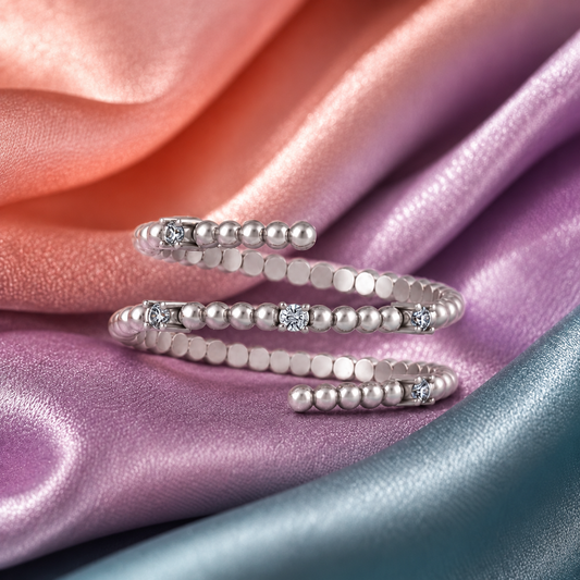 A silver-toned open bangle bracelet decorated with rows of small pearls and square-cut clear stones, arranged on draped satin fabric in pink, purple, and blue tones.