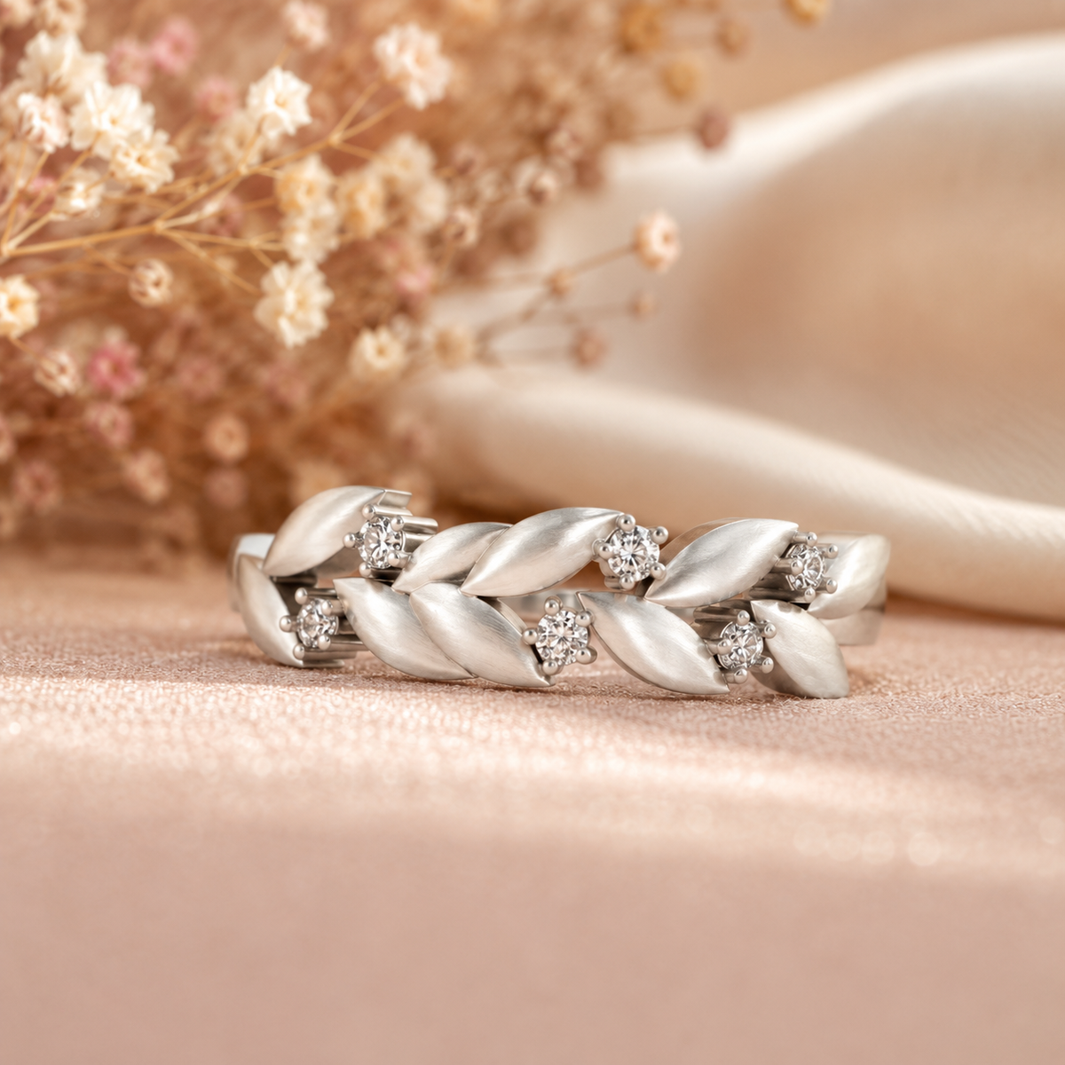 A silver-toned leaf-patterned ring with small clear gemstones, displayed on a soft beige surface with dried flowers and a satin ribbon in the background.