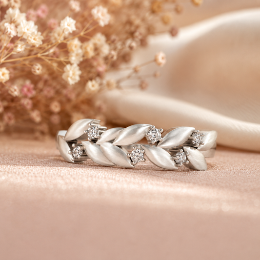 A silver-toned leaf-patterned ring with small clear gemstones, displayed on a soft beige surface with dried flowers and a satin ribbon in the background.