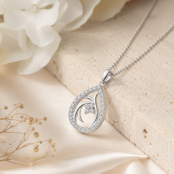 A silver teardrop diamond pendant necklace with a pavé-set circular border and a central curved openwork design, shown on a cream satin fabric with a textured stone slab and white flowers in the background.