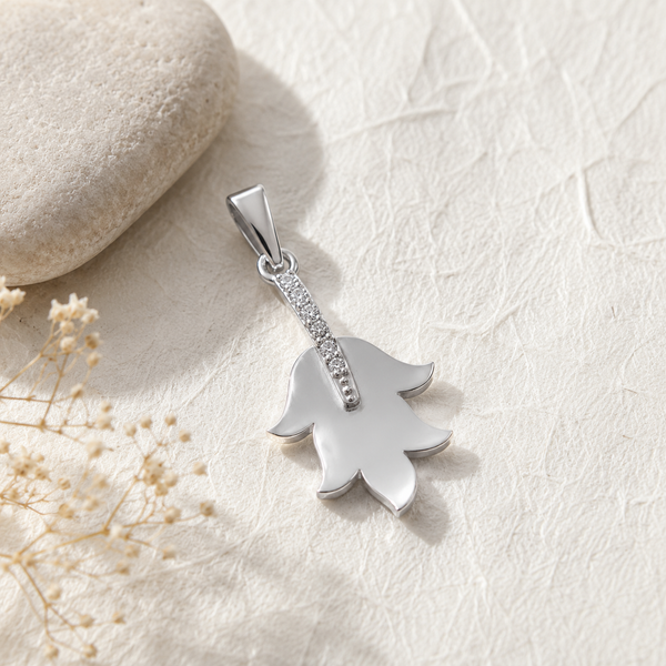 A silver diamond pendant shaped like a tulip with a small row of clear diamonds along the stem, photographed on a textured cream surface with a stone and dried flowers as props.
