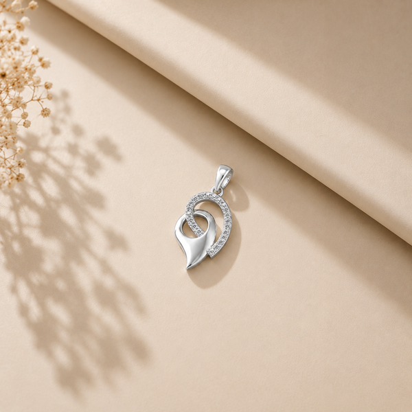 A silver pendant with a stylized double-heart design, one heart featuring a border of small clear diamond , displayed on a beige textured surface with soft shadows and dried flowers in the corner.