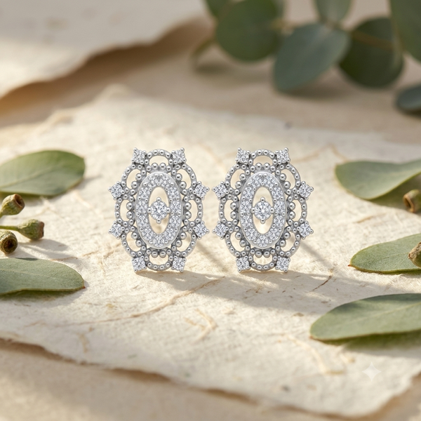 A pair of silver-toned filigree stud diamond earrings adorned with clear pavé diamonds , displayed on a textured beige stone surface with soft green eucalyptus leaves.