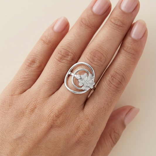 A silver ring with a flower and circular swirl design, shown worn on a hand with light pink manicured nails against a warm beige background.