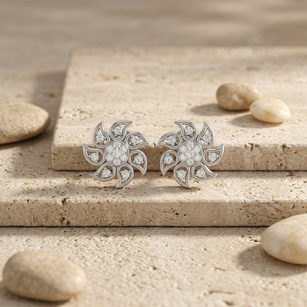 A pair of silver-toned flower-shaped stud diamonds earrings set with clear diamonds, displayed on a beige stone slab with small decorative pebbles around it.