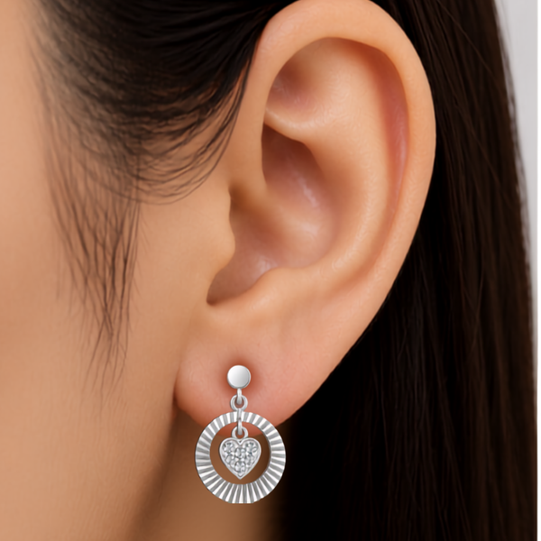 A close-up of a model's ear wearing a silver dangle diamond  earring with a sunburst outer ring, a small heart-shaped crystal center, and a round stud above.