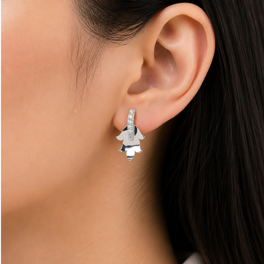 A close-up of a model's ear wearing a silver huggie hoop diamond earring with a dangling silver flower charm.