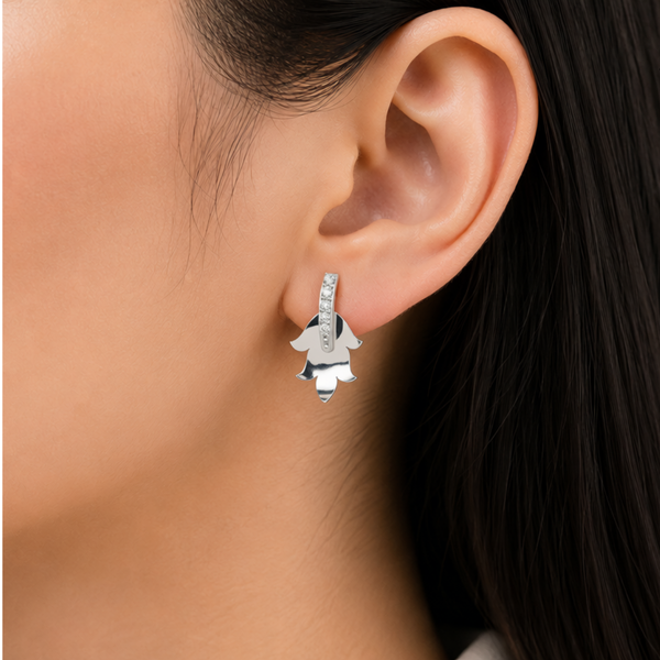 A close-up of a model's ear wearing a silver huggie hoop diamond earring with a dangling silver flower charm.