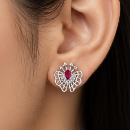A close-up of a person's ear wearing a butterfly-shaped stud earring. The earring features silver-toned butterfly wings with a central teardrop-shaped pink gemstone and small clear Diamond at the top.