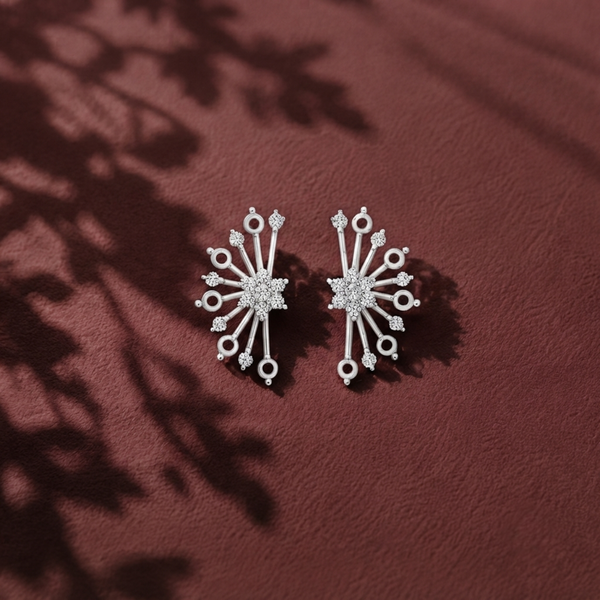 A pair of silver-toned starburst stud earrings with clear Diamonds, displayed on a textured dark red surface with leaf shadows.