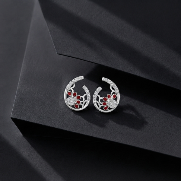 A pair of silver crescent-shaped hoop earrings with red floral inlay and clear Diamond accents, displayed on a dark textured geometric surface.