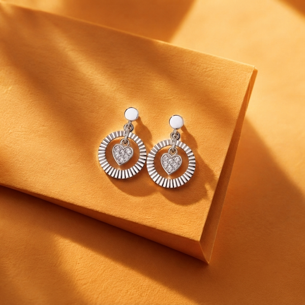 A pair of silver dangle diamond earrings with sunburst circular designs and heart-shaped center details, displayed on a textured orange surface with soft shadows.