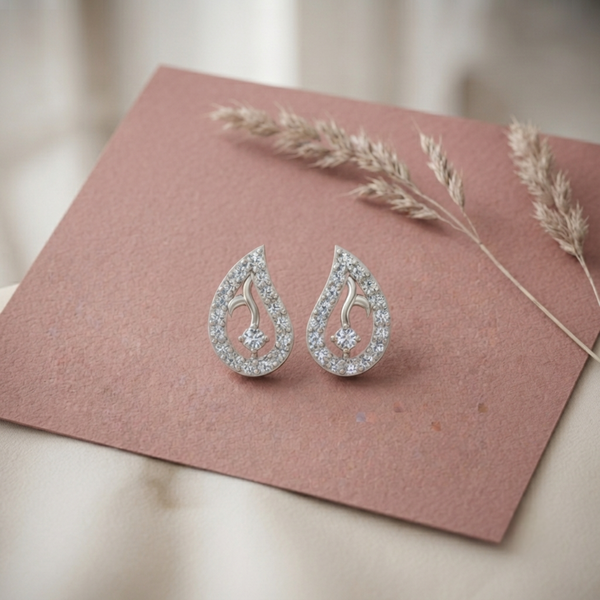 A pair of silver teardrop-shaped stud diamond earrings encrusted with clear diamond , displayed on a dusty rose textured card with dried grass stems.