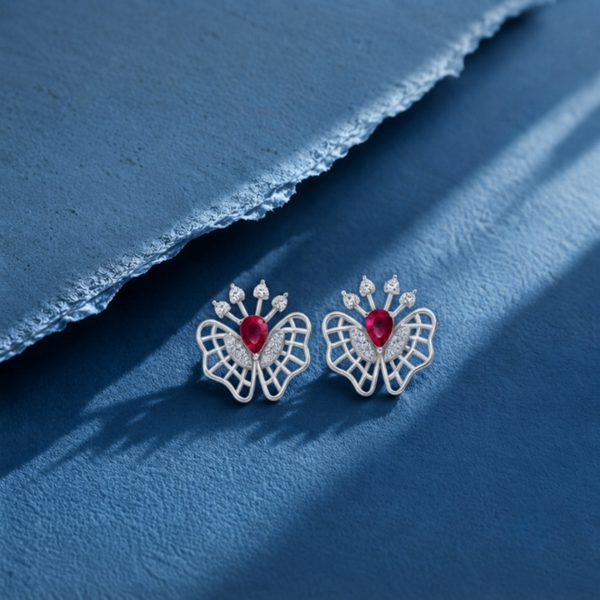 A pair of silver butterfly stud earrings with a central red heart-shaped gemstone and small clear Diamonds along the wings, displayed on a textured blue surface with a torn paper accent.