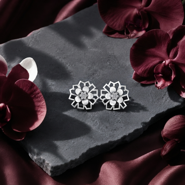 A pair of silver-toned geometric flower stud diamond earrings displayed on a dark gray slate slab, surrounded by deep burgundy orchid flowers and draped satin fabric.