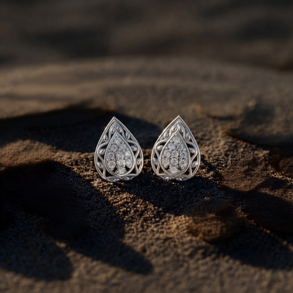 A pair of silver filigree teardrop stud  diamonds earrings with clear diamonds, resting on dark sand in warm, low-angle sunlight.
