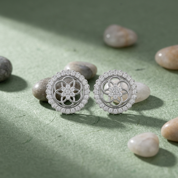 A pair of round silver-toned earrings with intricate floral filigree centers and rows of small clear diamonds, displayed on a textured sage green surface with scattered smooth diamonds.