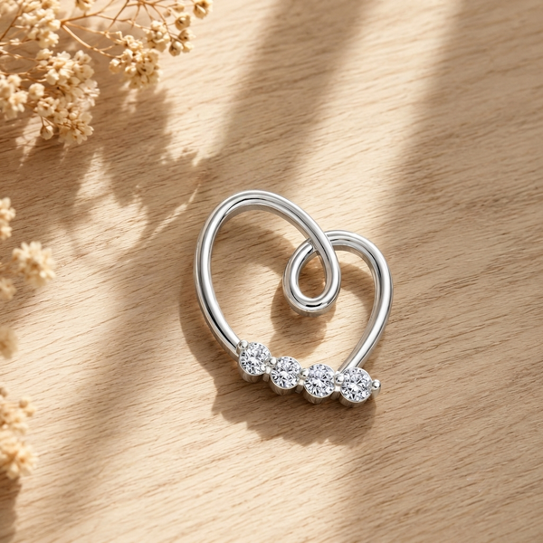 A silver heart-shaped pendant with a row of clear round-cut diamond along the lower curve, photographed on a light wood surface with soft dried flowers in the corner and dappled sunlight.