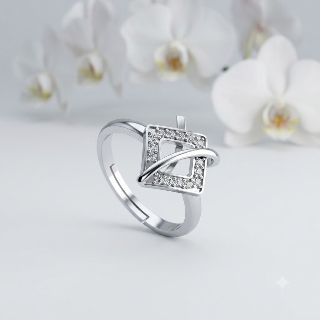 A silver-toned ring with a geometric square design set with small clear stones, photographed on a soft light gray surface with blurred white flowers in the background.