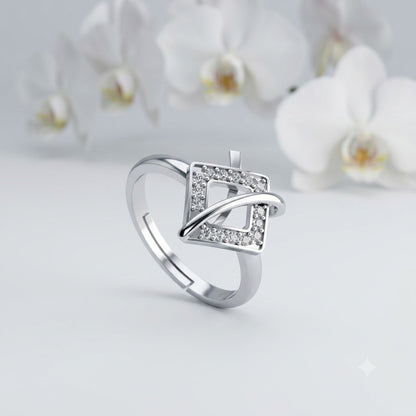 A silver-toned ring with a geometric square design set with small clear stones, photographed on a soft light gray surface with blurred white flowers in the background.